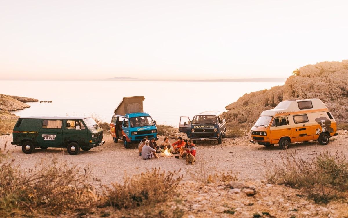 Mehrere Vans vor einer Meer-Kulisse, Menschen sitzen am Lagerfeuer
