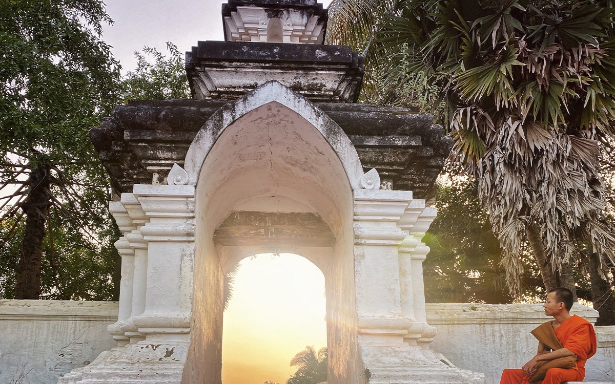 Laotischer Mönch sitzt bei Sonnenuntergang am Eingang des Tempels Wat Aham in Luang Prabang, Laos