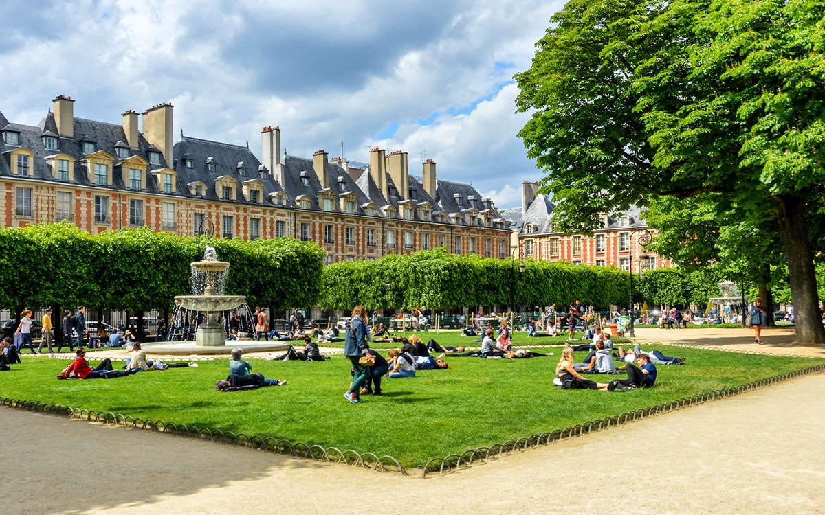 Ein Platz mit leuchtend grünem Gras, einem Brunnen und Menschen, die auf dem Rasen sitzen, umsäumt von einer Häuserreihe