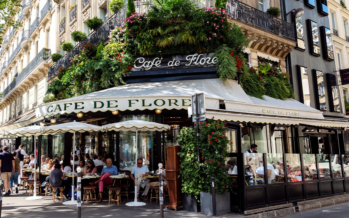 Ein Eckcafé in Paris, die Fassade ist mit Blumen bewachsen, Menschen sitzen im Außenbereich in der Sonne.
